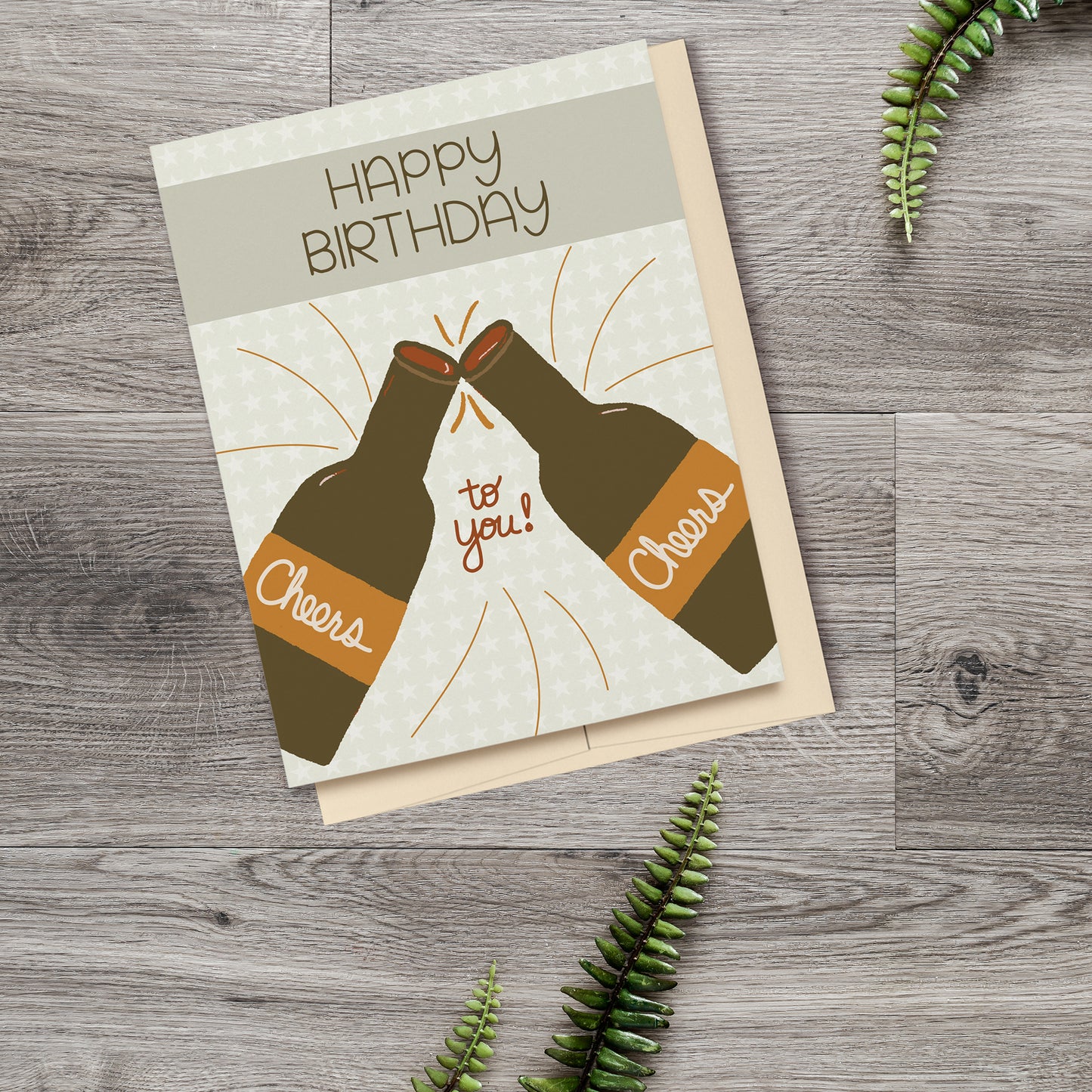 Greeting Card featuring two beer bottles clinking together with Cheers written on the labels, a banner at the top reading Happy Birthday and a star patterned background, displayed on a wooden surface with ferns.