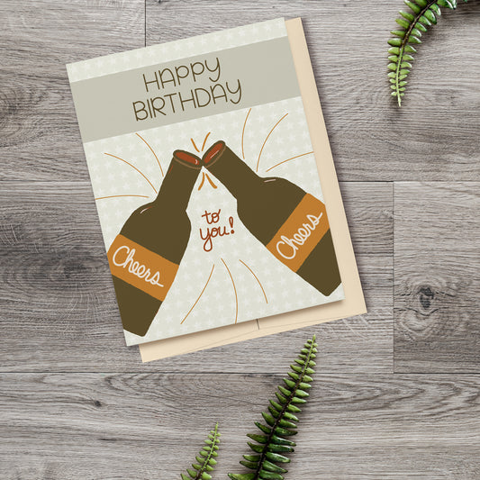 Greeting Card featuring two beer bottles clinking together with Cheers written on the labels, a banner at the top reading Happy Birthday and a star patterned background, displayed on a wooden surface with ferns.
