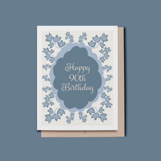 Birthday card featuring an abstract blue oval text frame with script text which reads, Happy 90th Birthday, surrounded by hand drawn blue botanical leaves with a cream and beige striped background.