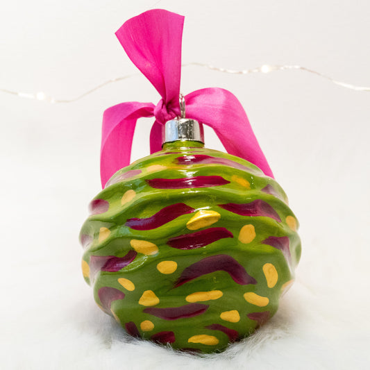 The Avery Hand Painted Ornament features a green gold base coat with rose violet and gold marks. Painted using fluid acrylic and acryla gouache paints. Displayed on white faux fur with fairy lights in the background.