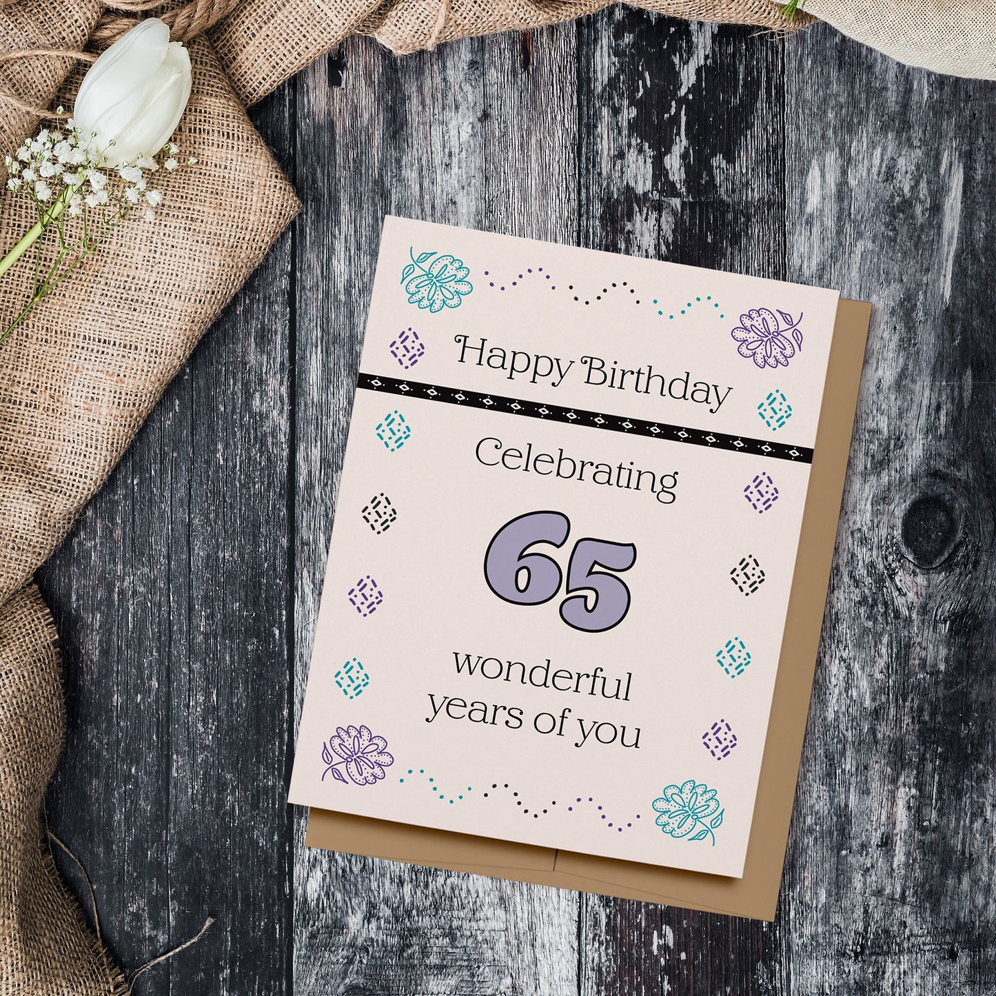 Pastel colored birthday card which reads, "Happy Birthday, Celebrating 65 wonderful years of you." Displayed on a wood background with burlap, a rose and baby's breath flowers.