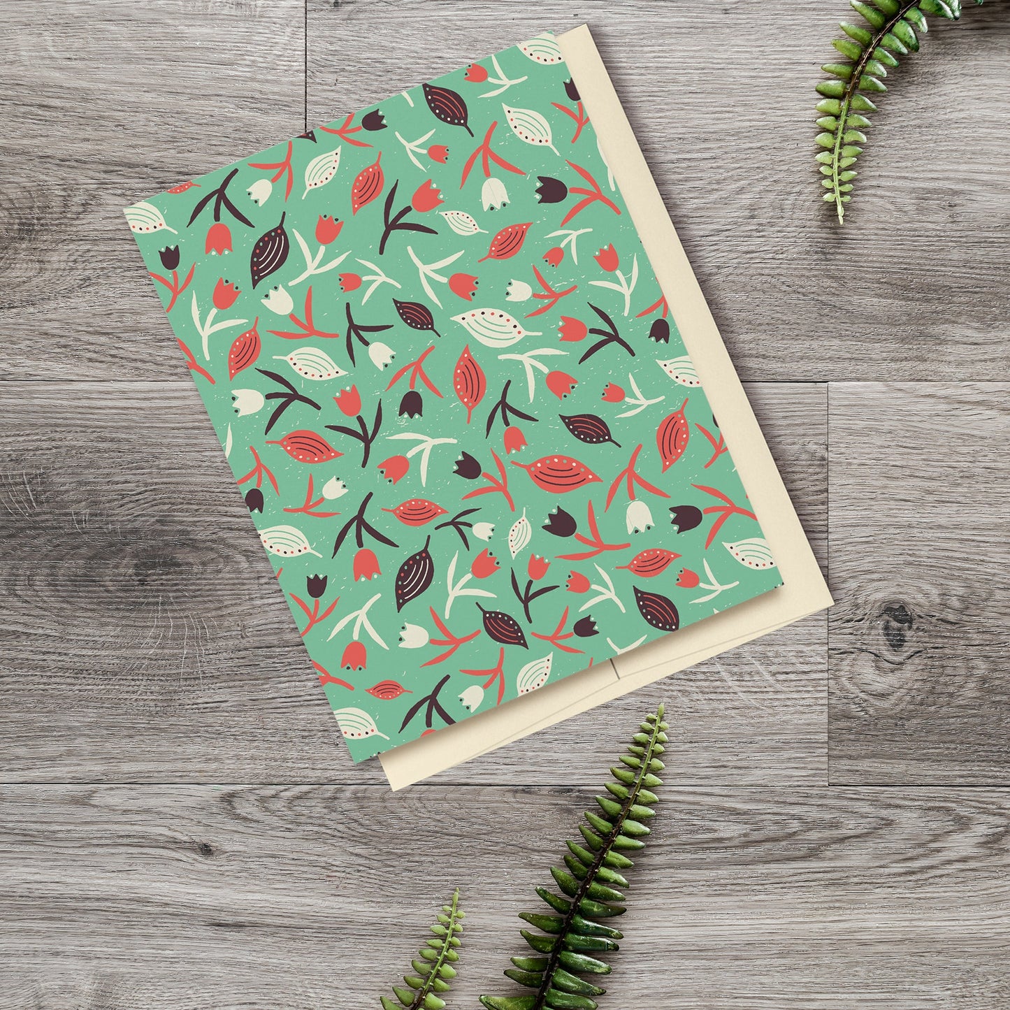 ulips & Leaves Seafoam Green & Coral Blank Card features a pattern with tulips and leaves in shades of cream, orange and brown on a seafoam green background. The pattern extends over the back of the card too. Displayed on a wooden background with ferns.