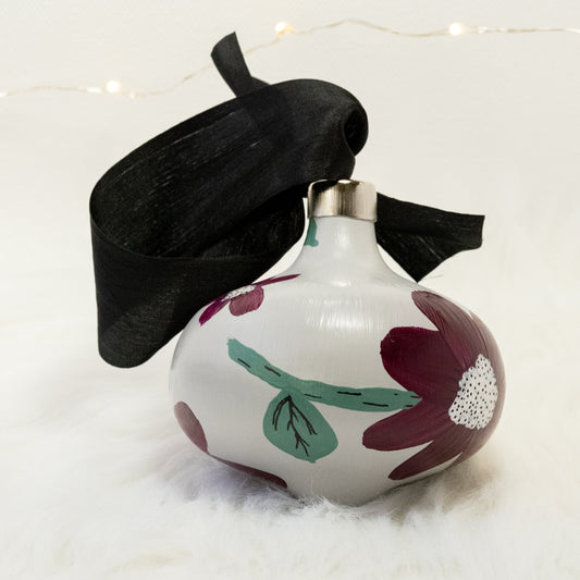 The Brenda Hand Painted Ornament features a light gray base coat, deep magenta flowers with sage green stems, white metallic and black accent details. Painted using acryla gouache paints. Displayed on white faux fur with fairy lights in the background.