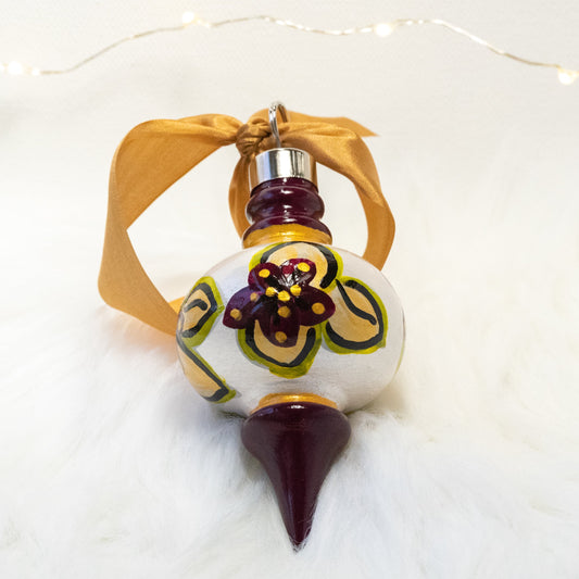 The Britta Hand Painted Ornament features a metallic white and deep magenta base coat, deep magenta flowers with gold and green gold leaves and gold and black accents. Painted using fluid acrylic and acryla gouache paints. Displayed on white faux fur with fairy lights in the background.