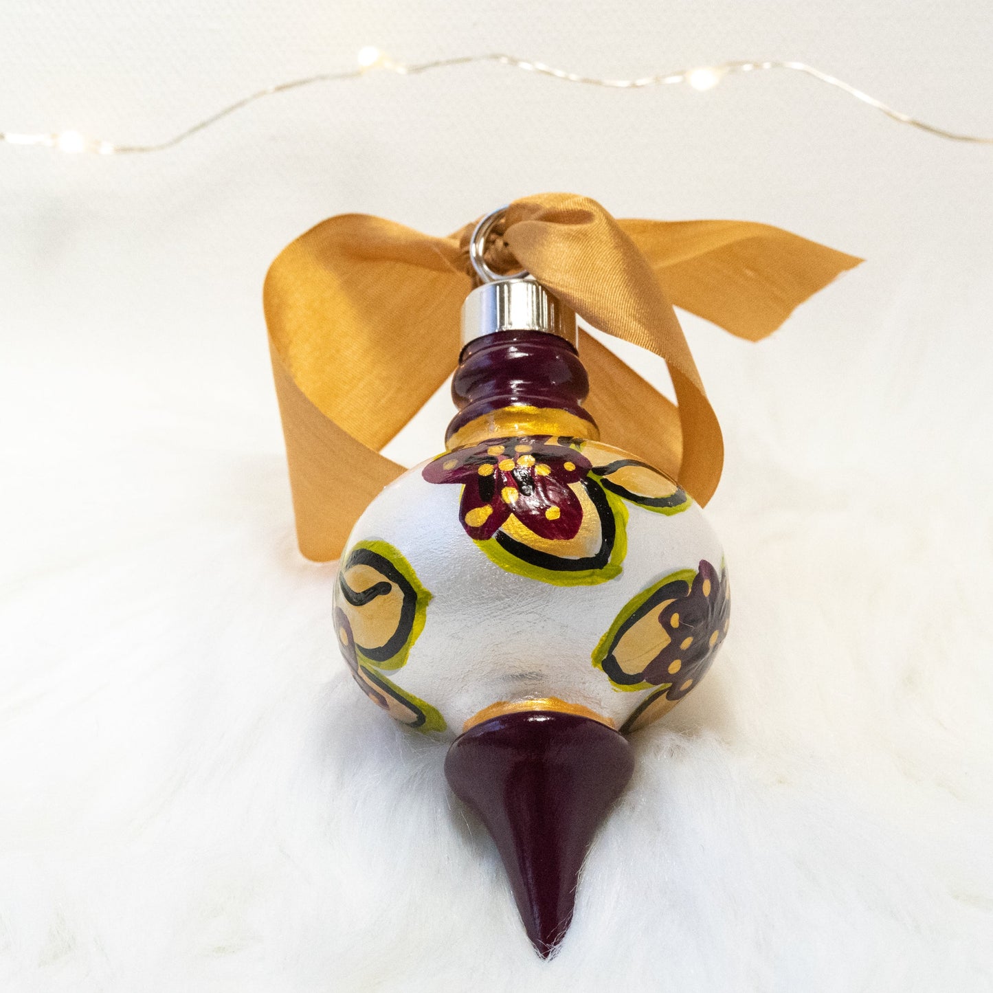 The Britta Hand Painted Ornament features a metallic white and deep magenta base coat, deep magenta flowers with gold and green gold leaves and gold and black accents. Painted using fluid acrylic and acryla gouache paints. Displayed on white faux fur with fairy lights in the background.