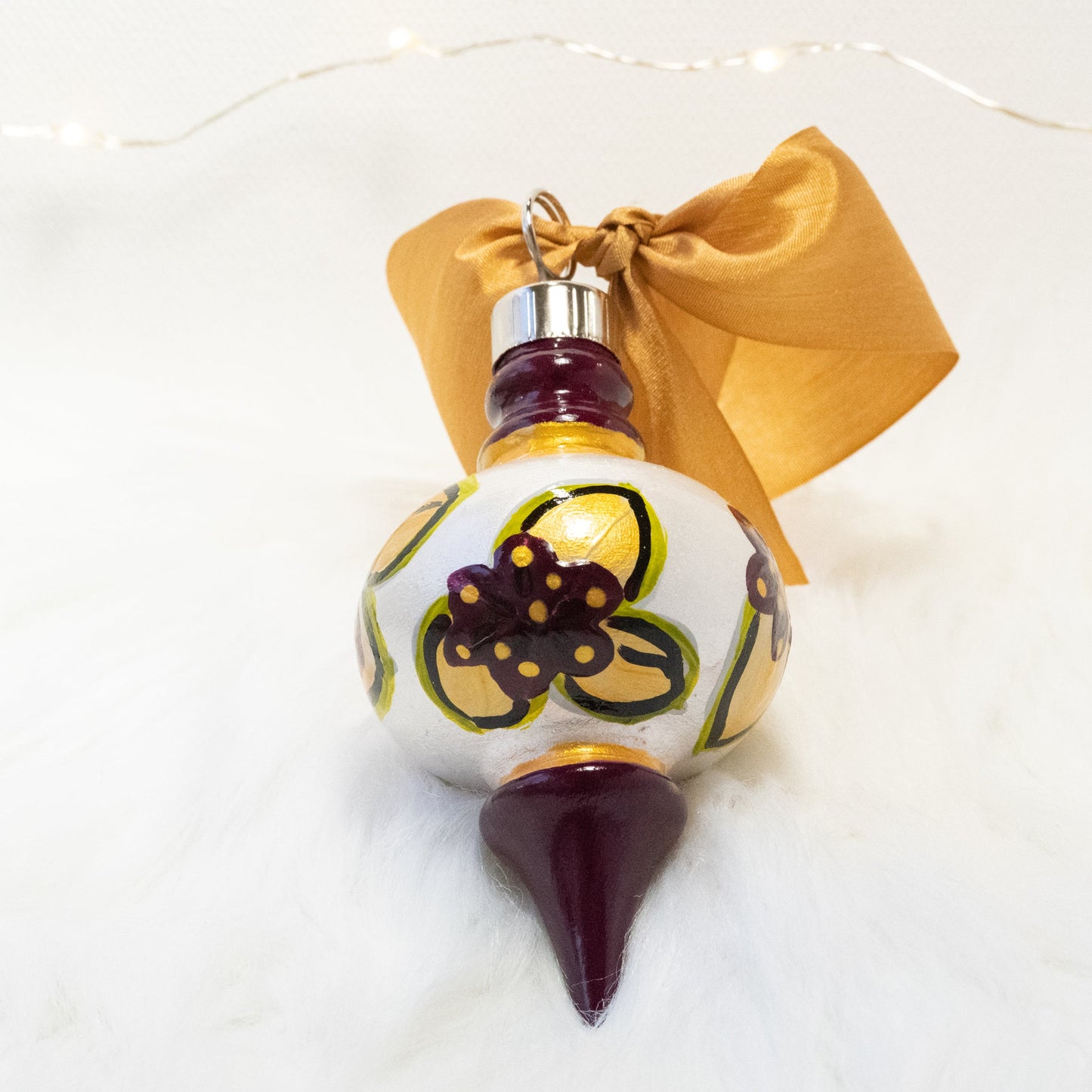 The Britta Hand Painted Ornament features a metallic white and deep magenta base coat, deep magenta flowers with gold and green gold leaves and gold and black accents. Painted using fluid acrylic and acryla gouache paints. Displayed on white faux fur with fairy lights in the background.