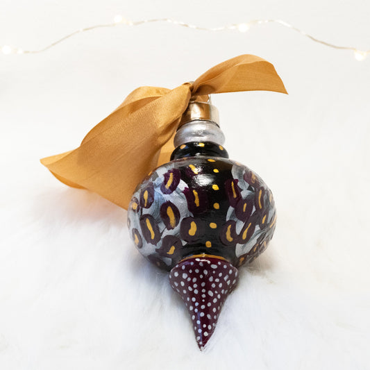 The Brook Lynn Hand Painted Ornament features a black and deep magenta base coat, silver, deep magenta and gold flowers with gold and silver accents. Painted using fluid acrylic and acryla gouache paints. Displayed on white faux fur with fairy lights in the background.