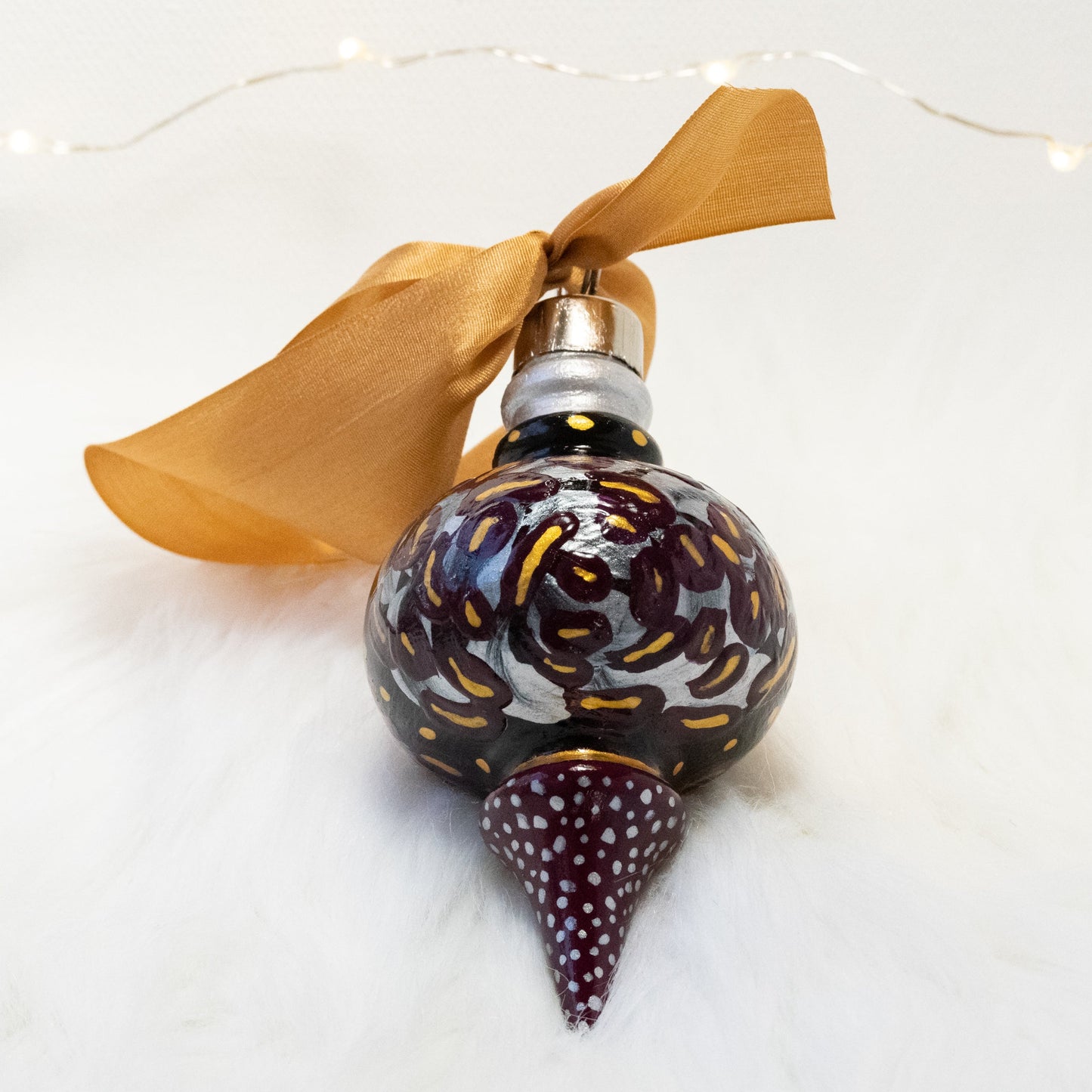 The Brook Lynn Hand Painted Ornament features a black and deep magenta base coat, silver, deep magenta and gold flowers with gold and silver accents. Painted using fluid acrylic and acryla gouache paints. Displayed on white faux fur with fairy lights in the background.