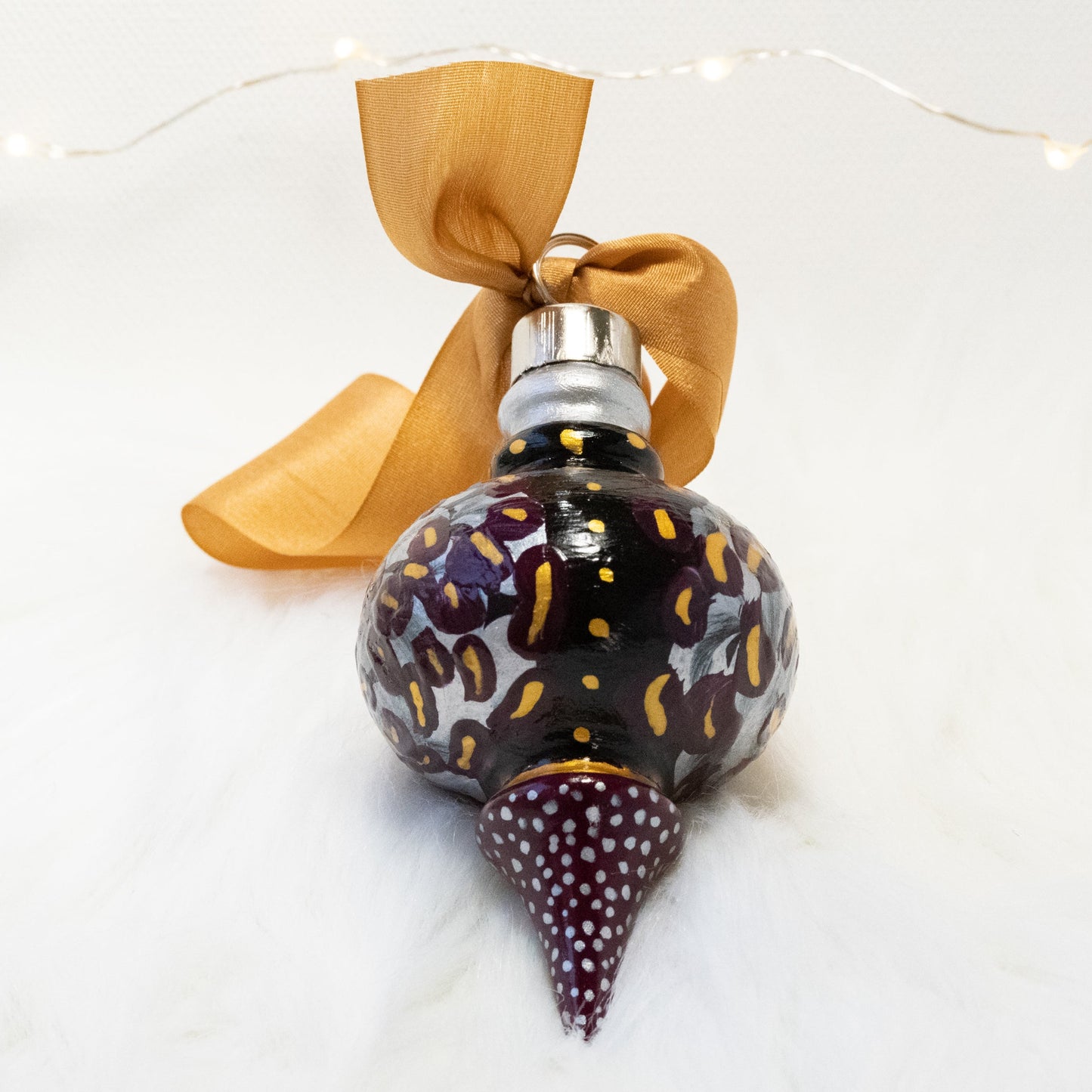 The Brook Lynn Hand Painted Ornament features a black and deep magenta base coat, silver, deep magenta and gold flowers with gold and silver accents. Painted using fluid acrylic and acryla gouache paints. Displayed on white faux fur with fairy lights in the background.