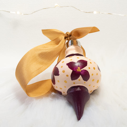 The Charlotte Hand Painted Ornament features a light peach and deep magenta base coat, deep magenta flowers with gold polka dots. Painted using fluid acrylic and acryla gouache paints. Displayed on white faux fur with fairy lights in the background.