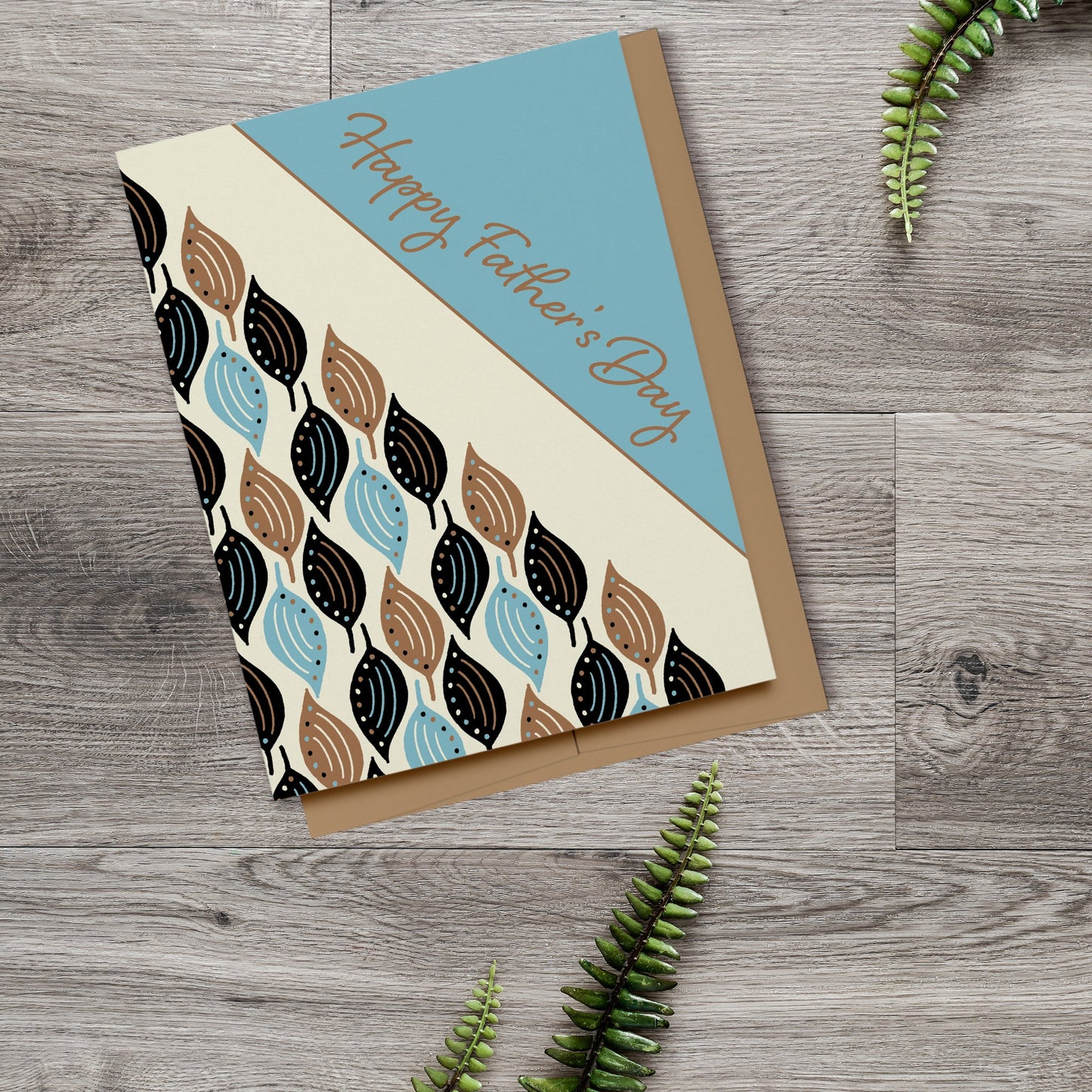 Happy Father's Day card with a pattern of blue, black and brown leaves on a cream background. Digitally hand drawn. Displayed on a wood background with fern leaves.