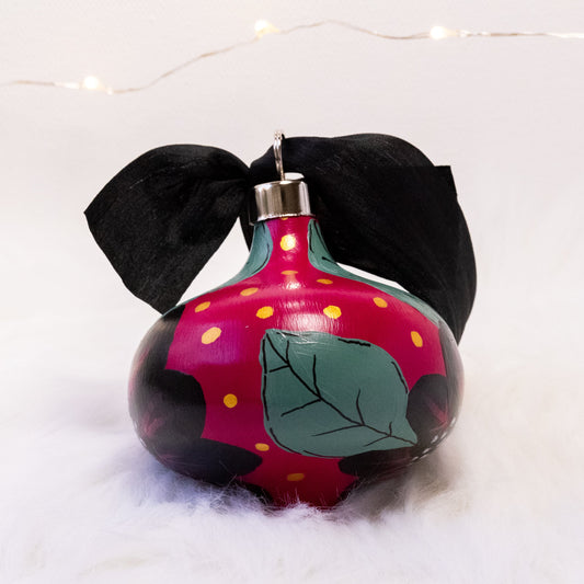 The Elizabeth Hand Painted Ornament features a rose violet base coat, black flowers with sage green leaves and black, rose violet, gold & metallic white accent details. Painted using acryla gouache paints. Displayed on white faux fur with fairy lights in the background.