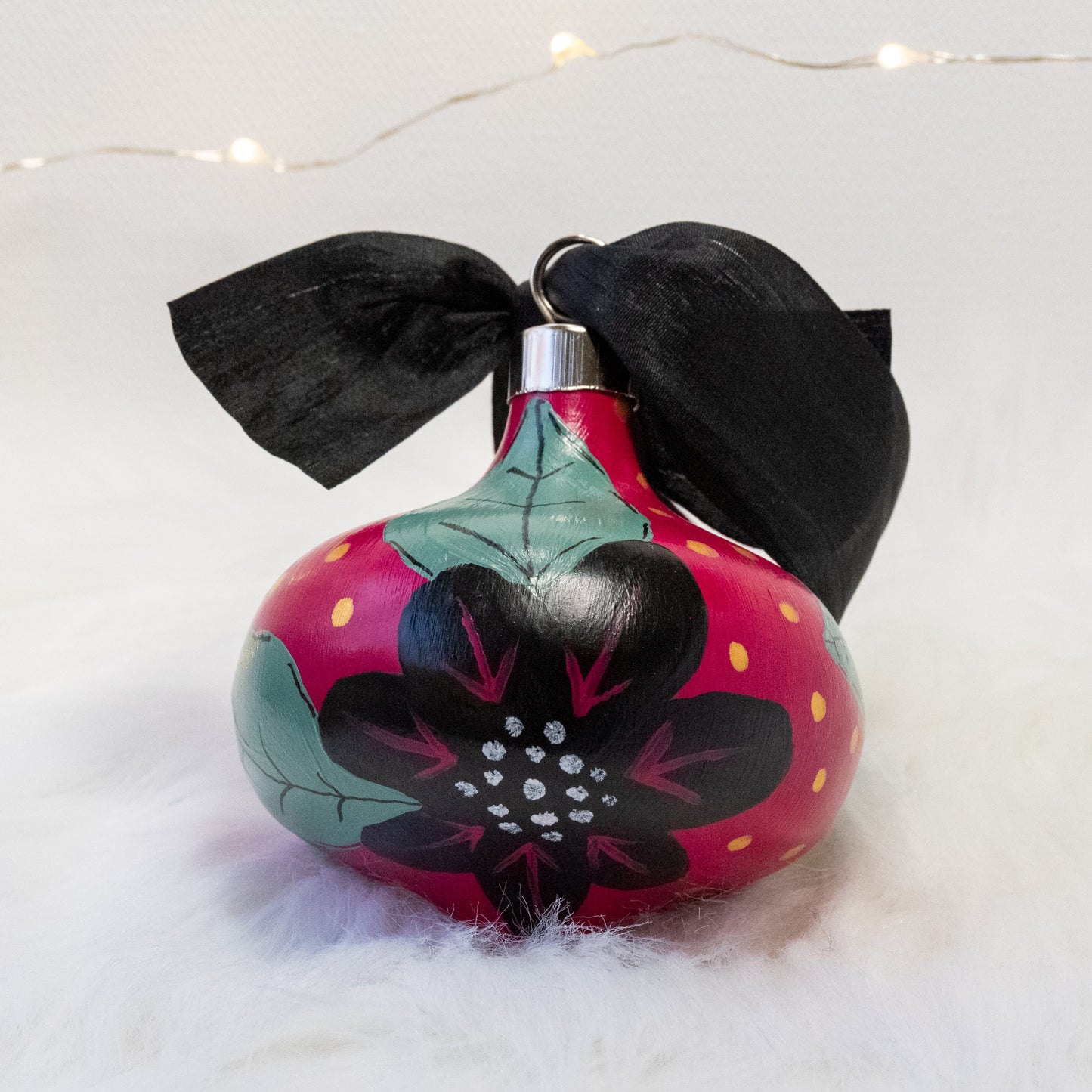The Elizabeth Hand Painted Ornament features a rose violet base coat, black flowers with sage green leaves and black, rose violet, gold & metallic white accent details. Painted using acryla gouache paints. Displayed on white faux fur with fairy lights in the background.