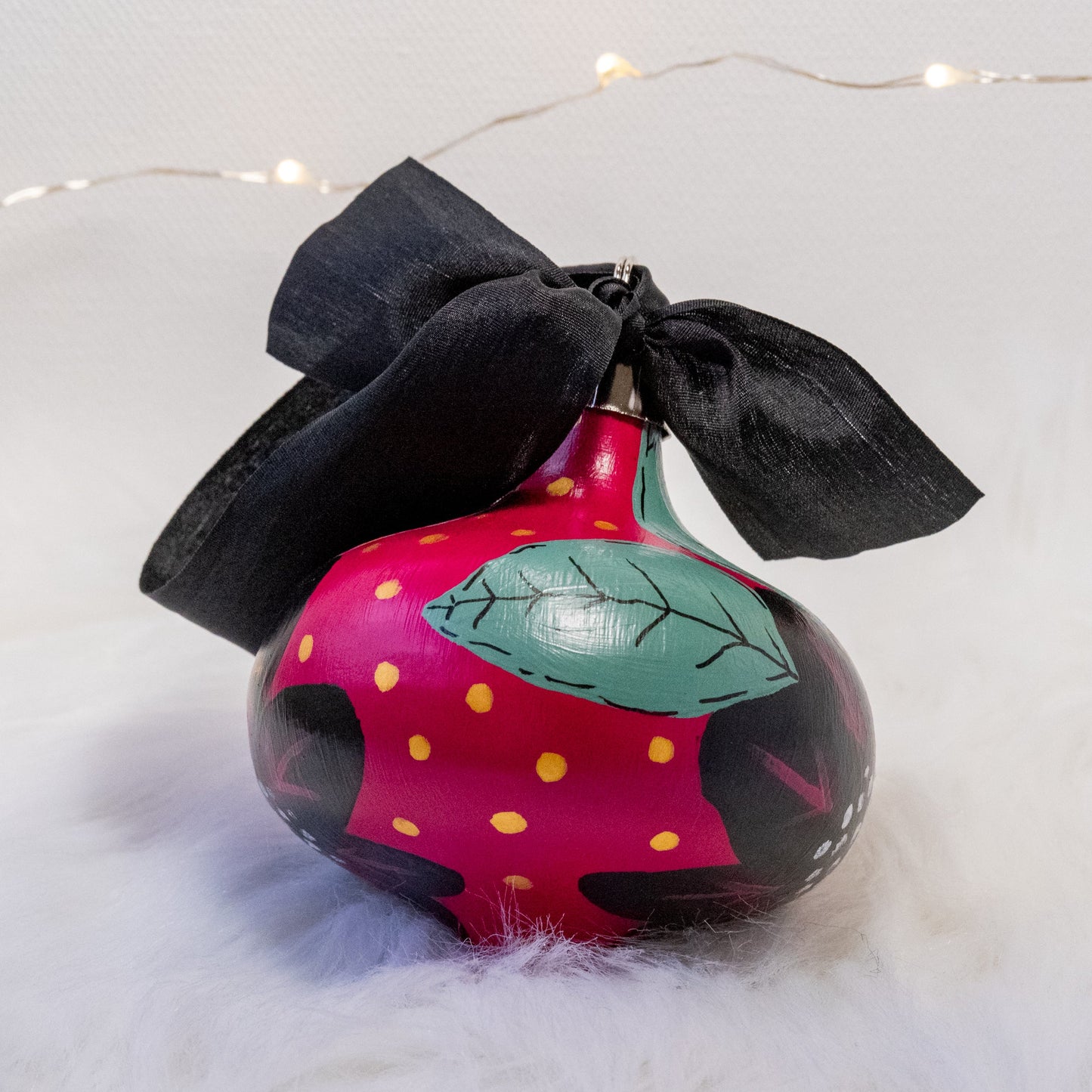 The Elizabeth Hand Painted Ornament features a rose violet base coat, black flowers with sage green leaves and black, rose violet, gold & metallic white accent details. Painted using acryla gouache paints. Displayed on white faux fur with fairy lights in the background.