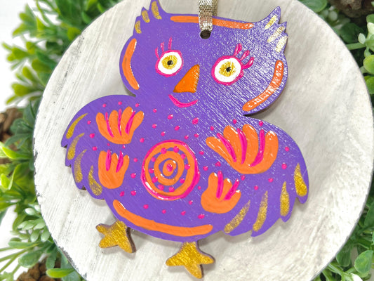 Purple Floral Owl Hand Painted Wood Ornament features a purple base with orange & pink flowers and pink dots and gold feathers & feet displayed on white wood surrounded by greenery and pine cones.