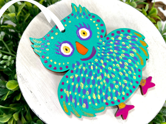 Aqua Owl Hand Painted Wood Ornament features an aqua base with yellow, purple & gold feathers, orange with fuchsia feet displayed on white wood surrounded by greenery and pine cones.