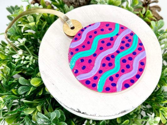 Fuchsia Round Hand Painted Wood Ornament features a fuchsia base with lavender & green wavy stripes, purple dots and a gold cap displayed on white wood surrounded by greenery and pine cones.