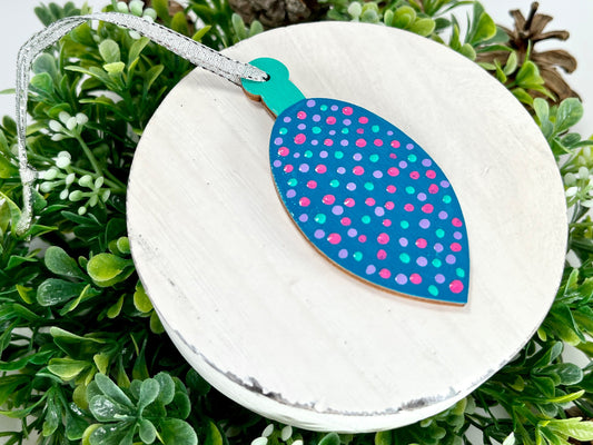 Blue Polka Dot Hand Painted Bulb Wood Ornament features a blue base with lavender, pink & aqua green polka dots and an aqua green cap displayed on white wood surrounded by greenery and pine cones.