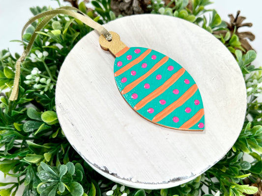 Aqua Green Striped Polka Dot Hand Painted Bulb Wood Ornament features an aqua green base with orange stripes and pink polka dots and a gold cap displayed on white wood surrounded by greenery and pine cones.