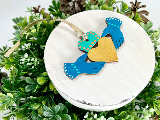 Deep Teal Irish Claddagh Hand Painted Wood Ornament features deep teal hands with green & white dots on the cuffs, an aqua green crown with orange dots and a gold heart displayed on white wood surrounded by greenery and pine cones.