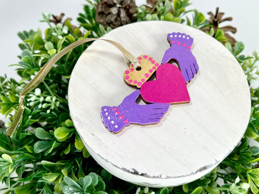 Purple Irish Claddagh Hand Painted Wood Ornament features purple hands with pink & white dots on the cuffs, a gold crown with pink dots and a pink heart displayed on white wood surrounded by greenery and pine cones.