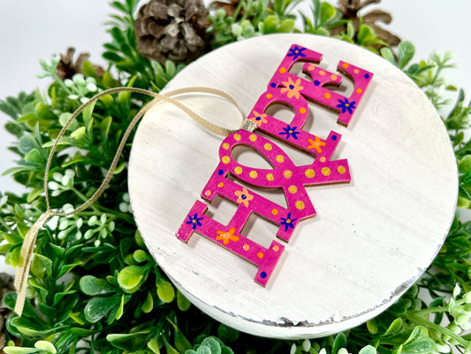 Fuchsia, Purple and Orange Hope Ribbon Hand Painted Wood Ornament features a fuchsia background with orange & purple flowers and dots and gold dots inside the ribbon displayed on white wood surrounded by greenery and pine cones.