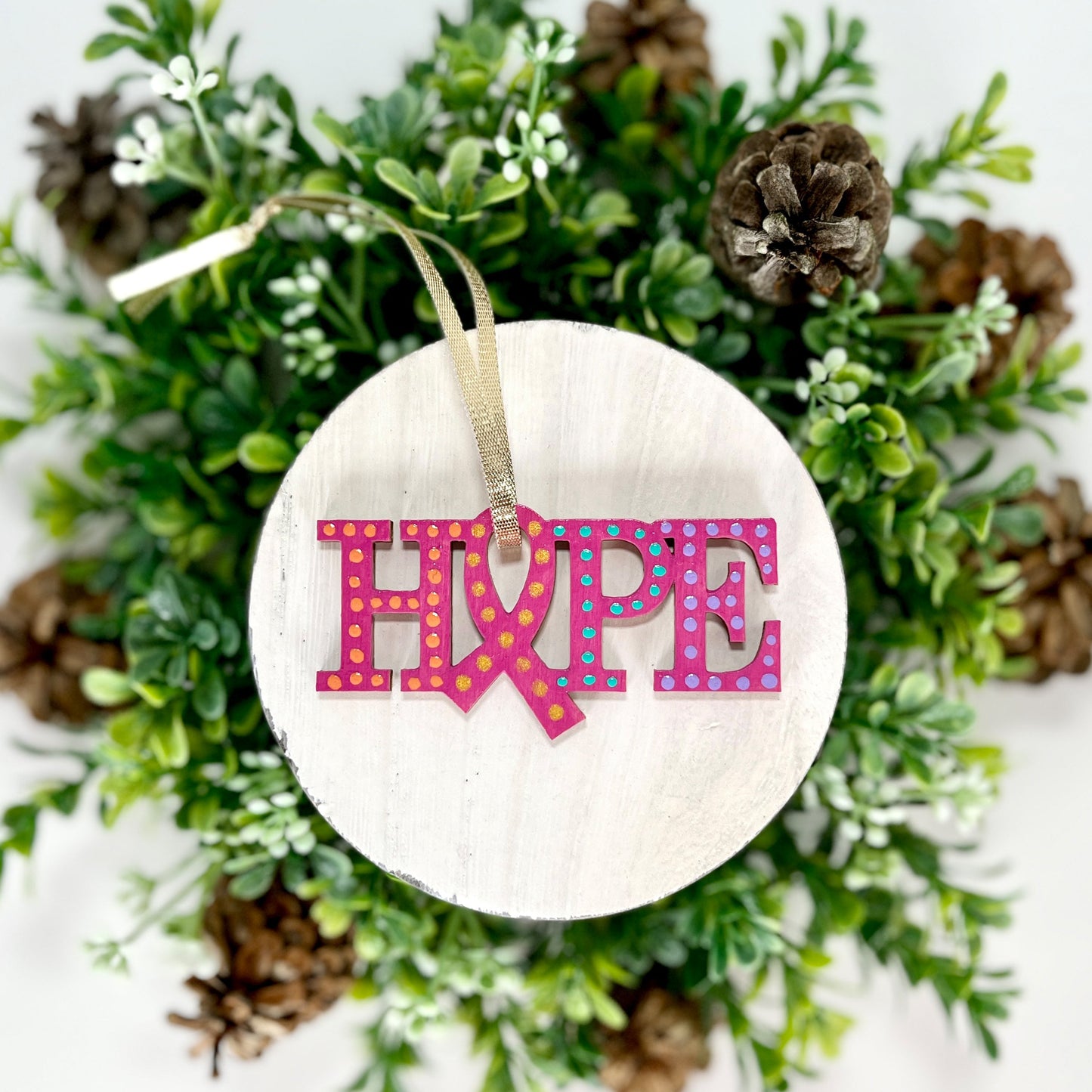 Fuchsia, Purple, Aqua and Orange Hope Ribbon Hand Painted Wood Ornament features a fuchsia background with orange, purple, aqua and gold dots. Includes a gold ribbon for hanging displayed on white wood surrounded by greenery and pine cones.