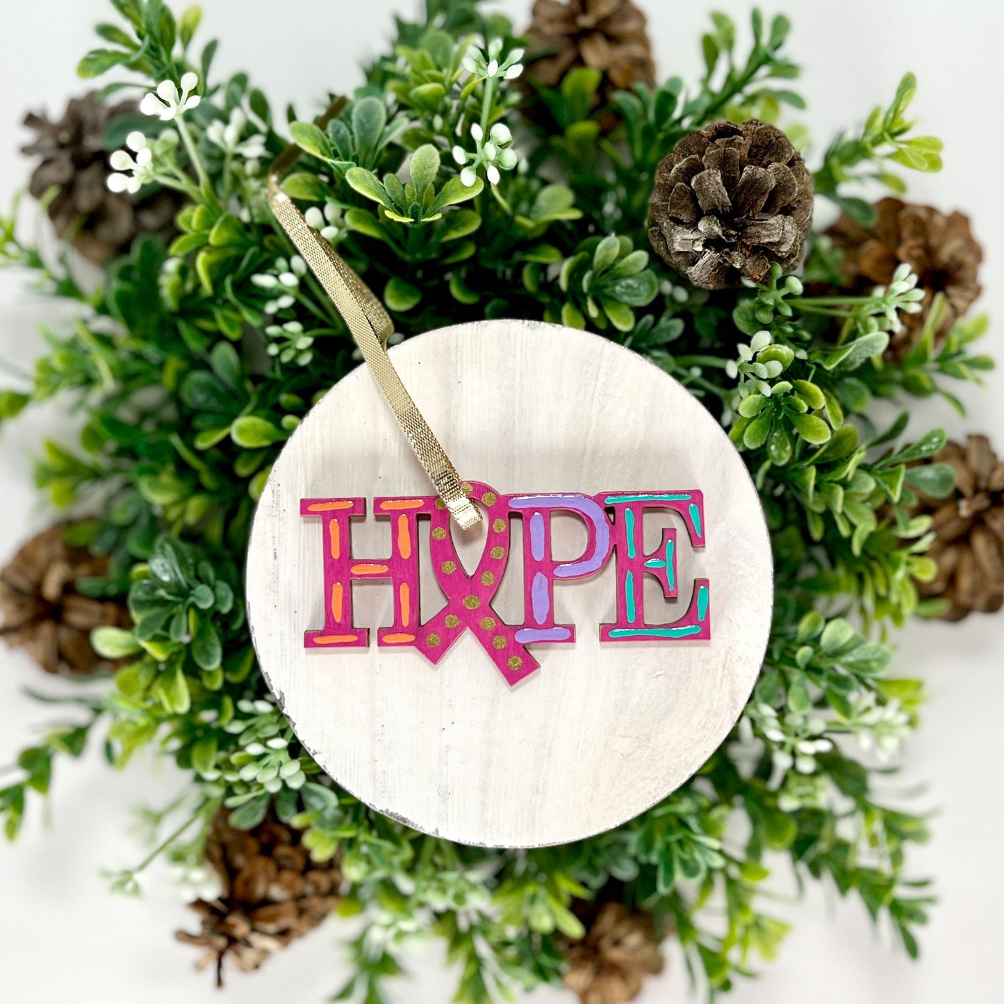 Fuchsia, Orange, Purple and Aqua Hope Ribbon Hand Painted Wood Ornament features a fuchsia background with orange, purple and aqua dashes with gold dots inside the ribbon displayed on white wood surrounded by greenery and pine cones.