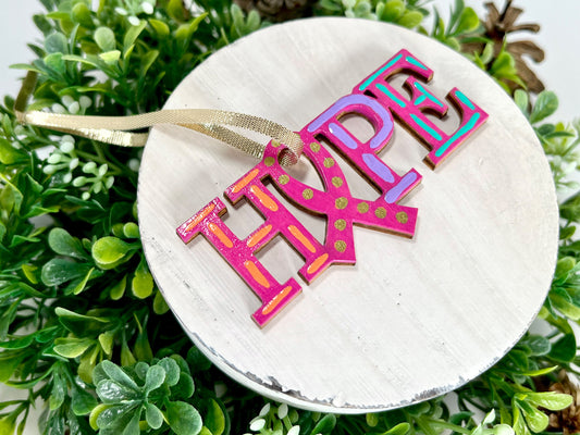 Fuchsia, Orange, Purple and Aqua Hope Ribbon Hand Painted Wood Ornament features a fuchsia background with orange, purple and aqua dashes with gold dots inside the ribbon displayed on white wood surrounded by greenery and pine cones.