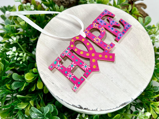 Fuchsia, White, Yellow and Purple Hope Ribbon Hand Painted Wood Ornament features a fuchsia background with white, yellow and purple flowers along with aqua, purple and blue dots and gold dots inside the ribbon displayed on white wood surrounded by greenery and pine cones.