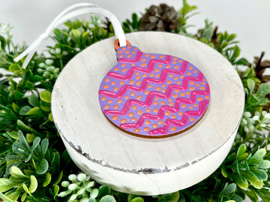 Purple, Pink & Orange Round Hand Painted Wood Ornament features purple with pink zig zags, orange polka dots and an orange cap displayed on white wood surrounded by greenery and pine cones.