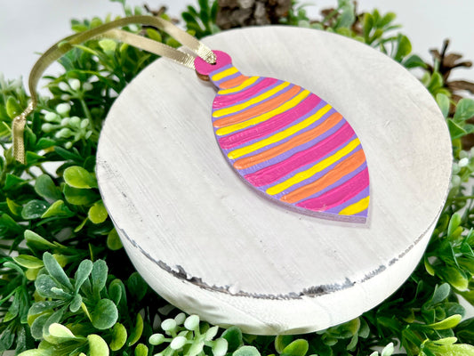 Purple Colorful Striped Bulb Hand Painted Wood Ornament features purple with yellow, pink & orange stripes displayed on white wood surrounded by greenery and pine cones.