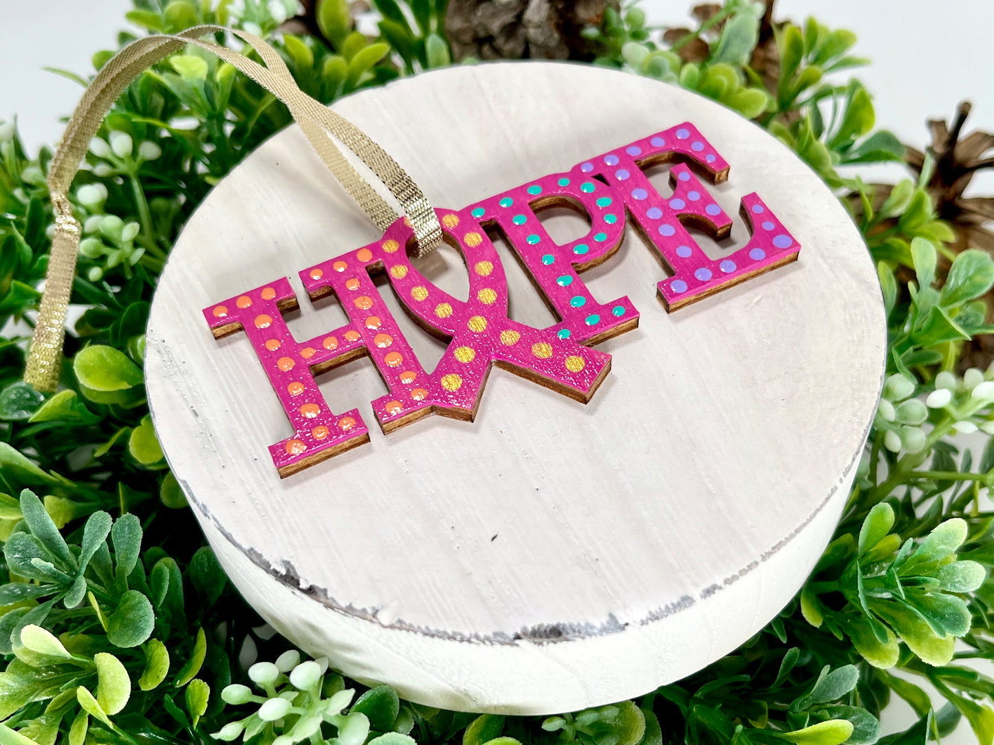 Fuchsia, Purple, Aqua and Orange Hope Ribbon Hand Painted Wood Ornament features a fuchsia background with orange, purple, aqua and gold dots. Includes a gold ribbon for hanging displayed on white wood surrounded by greenery and pine cones.