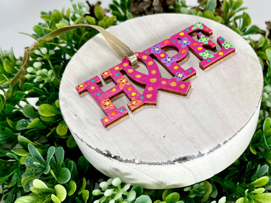 Fuchsia, Orange, Purple and Green Hope Ribbon Hand Painted Wood Ornament features a fuchsia background with orange, purple and green flowers along with aqua, yellow and orange dots and gold dots inside the ribbon displayed on white wood surrounded by greenery and pine cones.