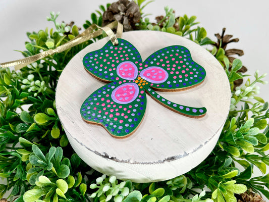 The Dark Green Shamrock Hand Painted Wood Ornament features a dark green base with a pink & purple flower, as well as pink & purple dots displayed on white wood surrounded by greenery and pine cones.