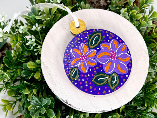 Deep Purple Floral Hand Painted Round Wood Ornament features a deep purple base with lavender, orange, pink & white flowers with green leaves and pink, gold & white dots with a gold cap displayed on white wood surrounded by greenery and pine cones.
