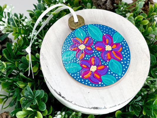 Blue Floral Hand Painted Round Wood Ornament features a blue base with lavender, pink & orange flowers with aqua green leaves, white polka dots and a gold cap displayed on white wood surrounded by greenery and pine cones.
