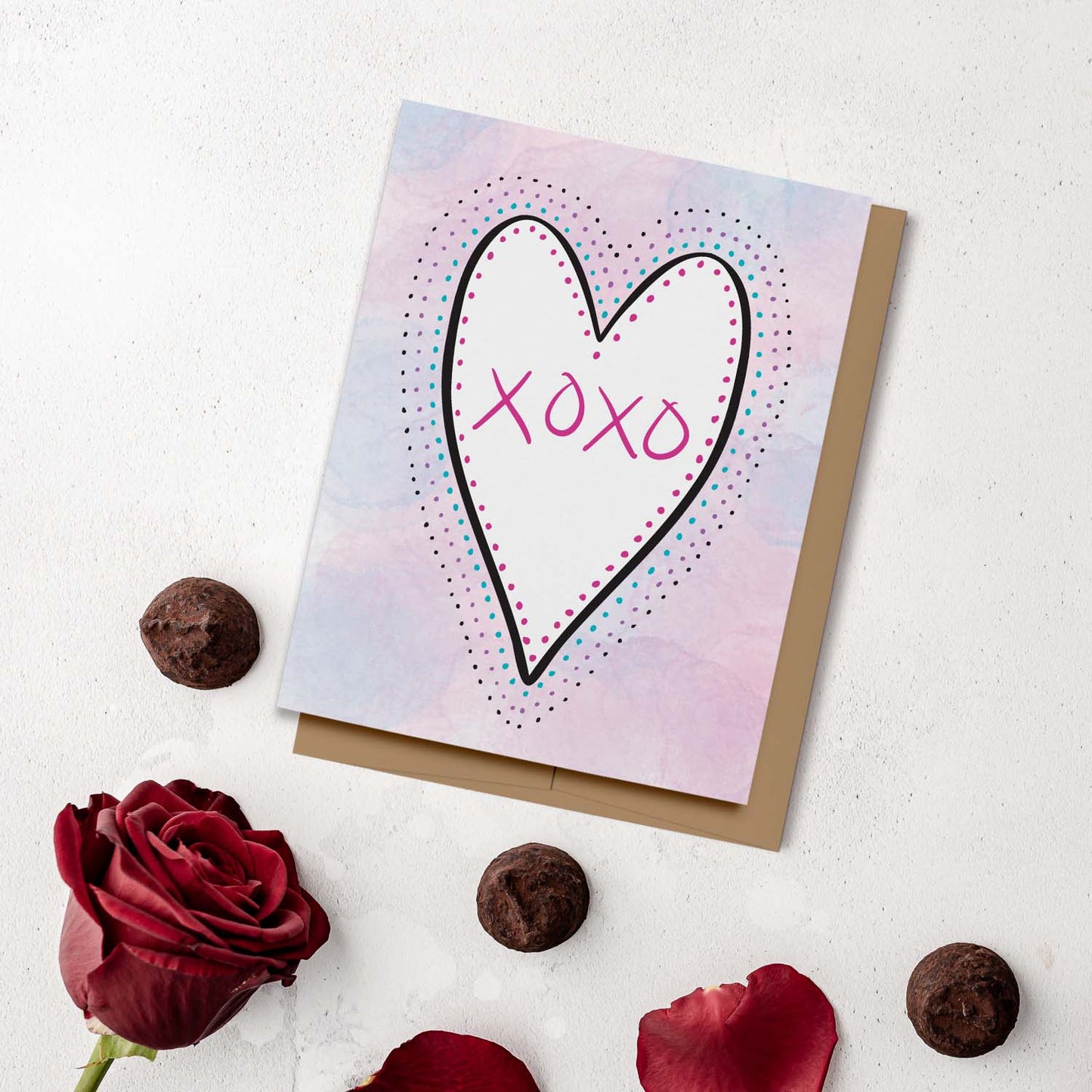 Love card featuring a white heart with hand lettered XOXO outlined with polka dots on a pink, purple and blue watercolor background. Displayed on a white cement background with red roses and chocolate truffles.