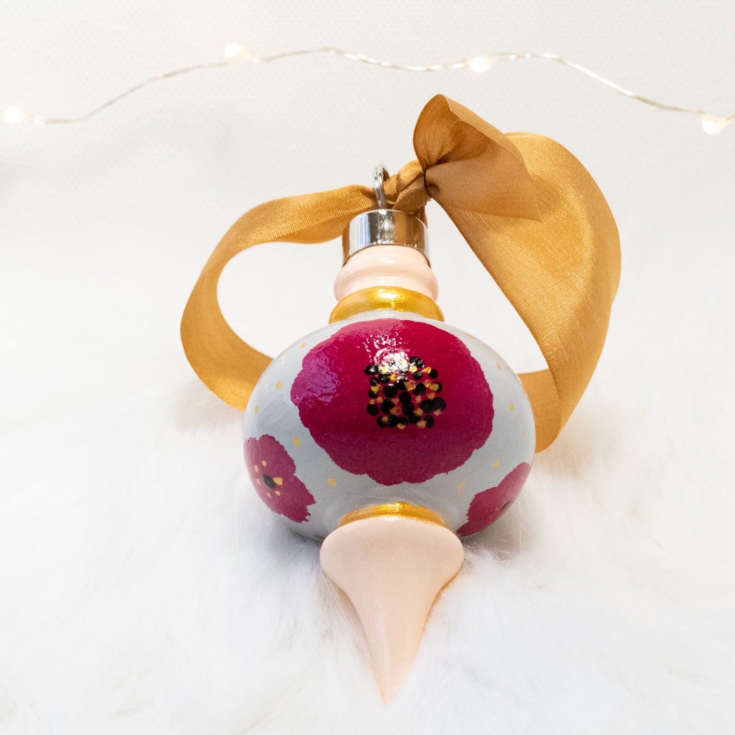 The Lulu Hand Painted Ornament features a light gray and peach base coat, rose violet flowers with gold polka dots and black and gold accents. Painted using fluid acrylic and acryla gouache paints. Displayed on white faux fur with fairy lights in the background.