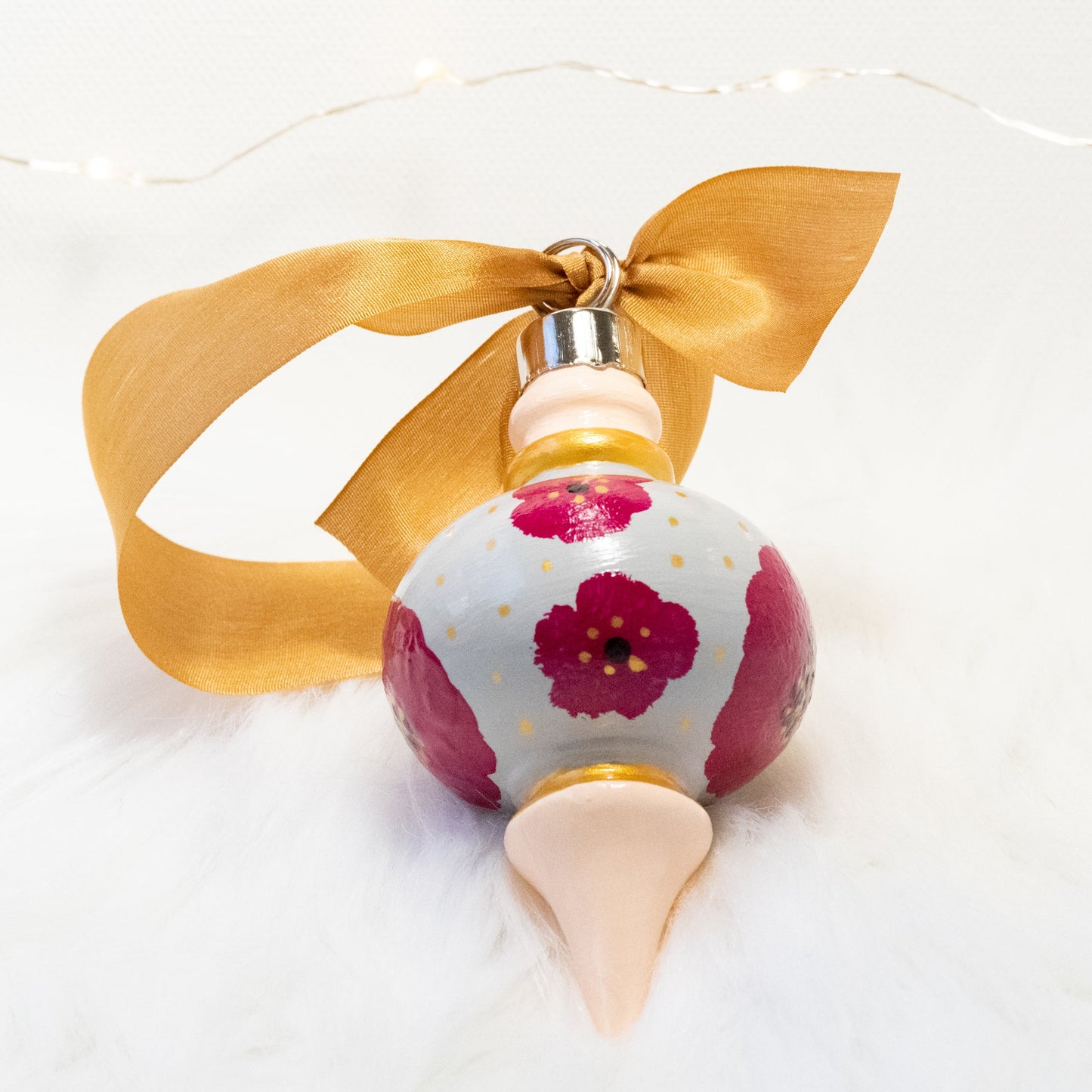 The Lulu Hand Painted Ornament features a light gray and peach base coat, rose violet flowers with gold polka dots and black and gold accents. Painted using fluid acrylic and acryla gouache paints. Displayed on white faux fur with fairy lights in the background.