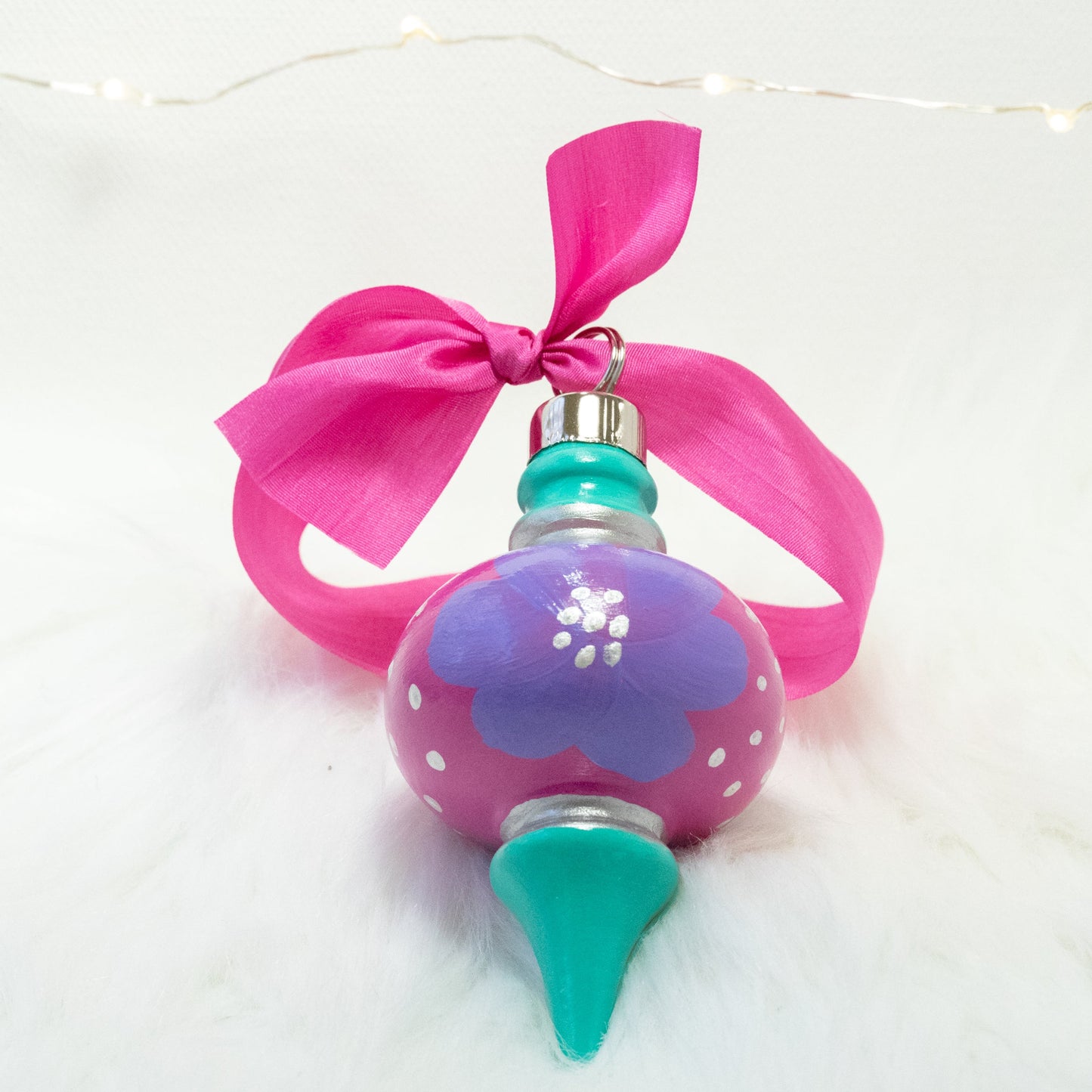 The Maxie Hand Painted Ornament features a fuchsia and bright aqua green base coat, lilac purple flowers with silver accents. Painted using fluid acrylic and acryla gouache paints. Displayed on white faux fur with fairy lights in the background.