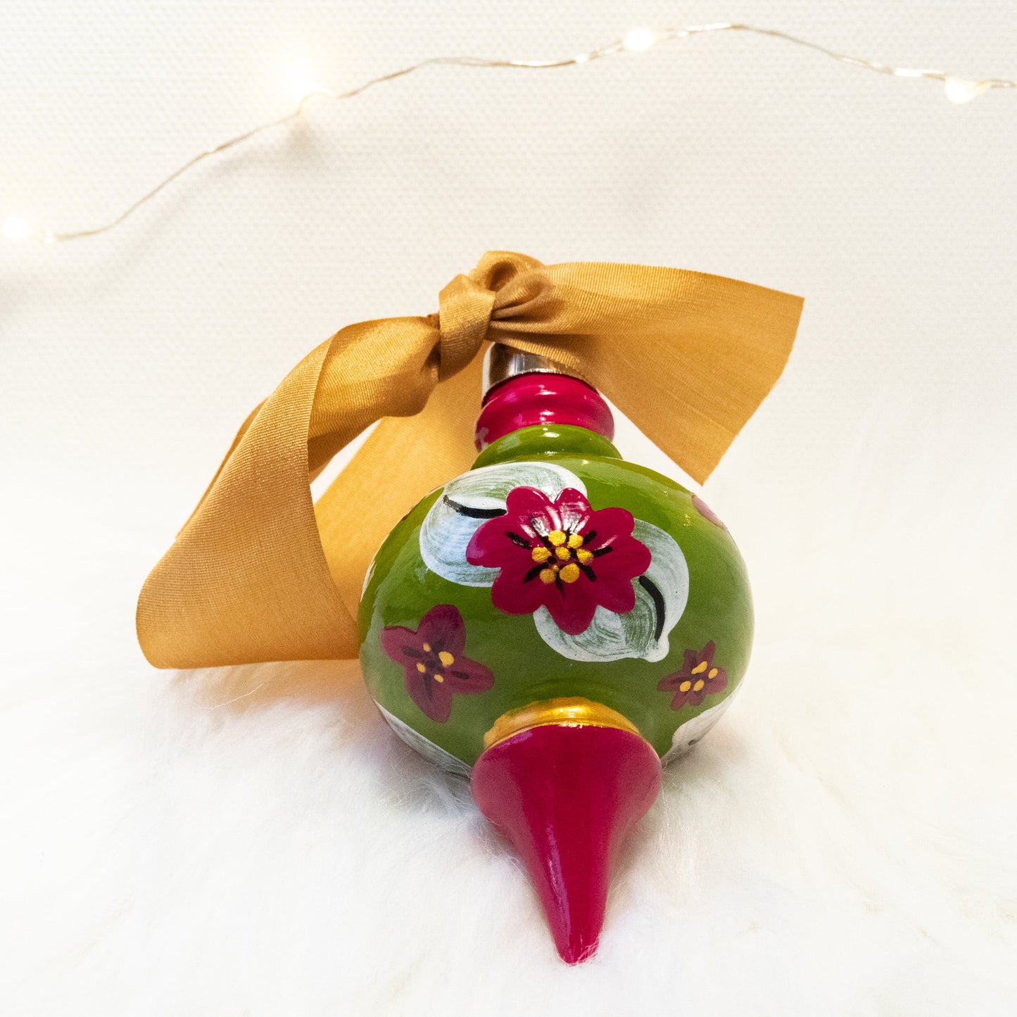 The Nina Hand Painted Ornament features a green gold and rose violet base coat, rose violet flowers with metallic white leaves with black and gold accents. Painted using fluid acrylic and acryla gouache paints. Displayed on white faux fur with fairy lights in the background.
