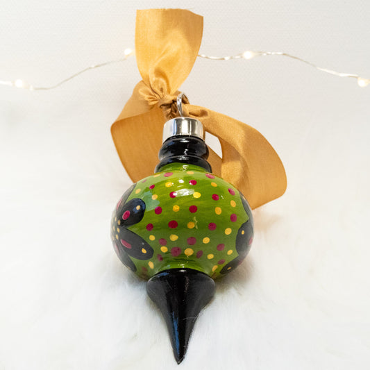 The Sam Hand Painted Ornament features a green gold and black base coat, black flowers with rose violet and gold accents and rose violet and gold polka dots. Painted using fluid acrylic and acryla gouache paints. Displayed on white faux fur with fairy lights in the background.