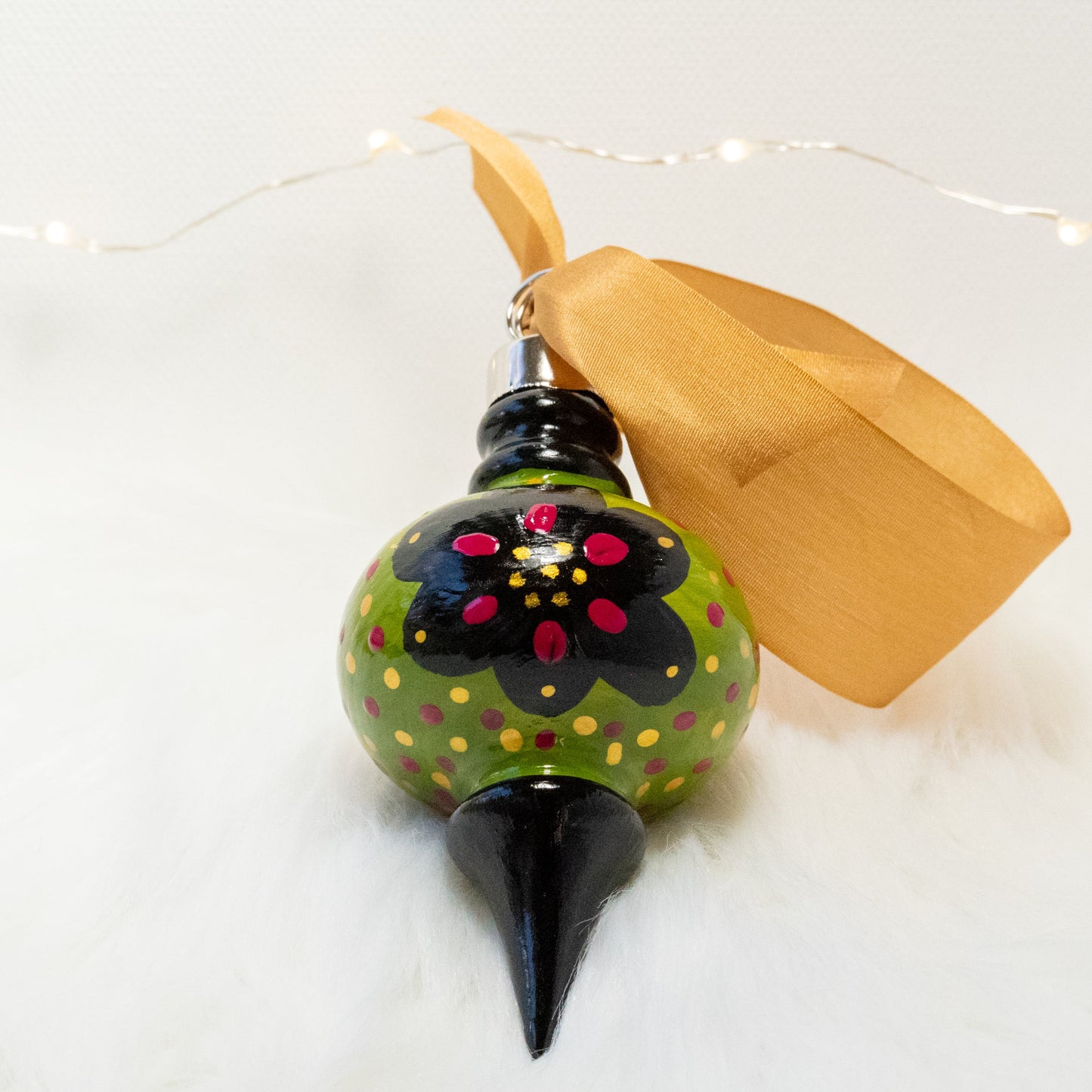 The Sam Hand Painted Ornament features a green gold and black base coat, black flowers with rose violet and gold accents and rose violet and gold polka dots. Painted using fluid acrylic and acryla gouache paints. Displayed on white faux fur with fairy lights in the background.