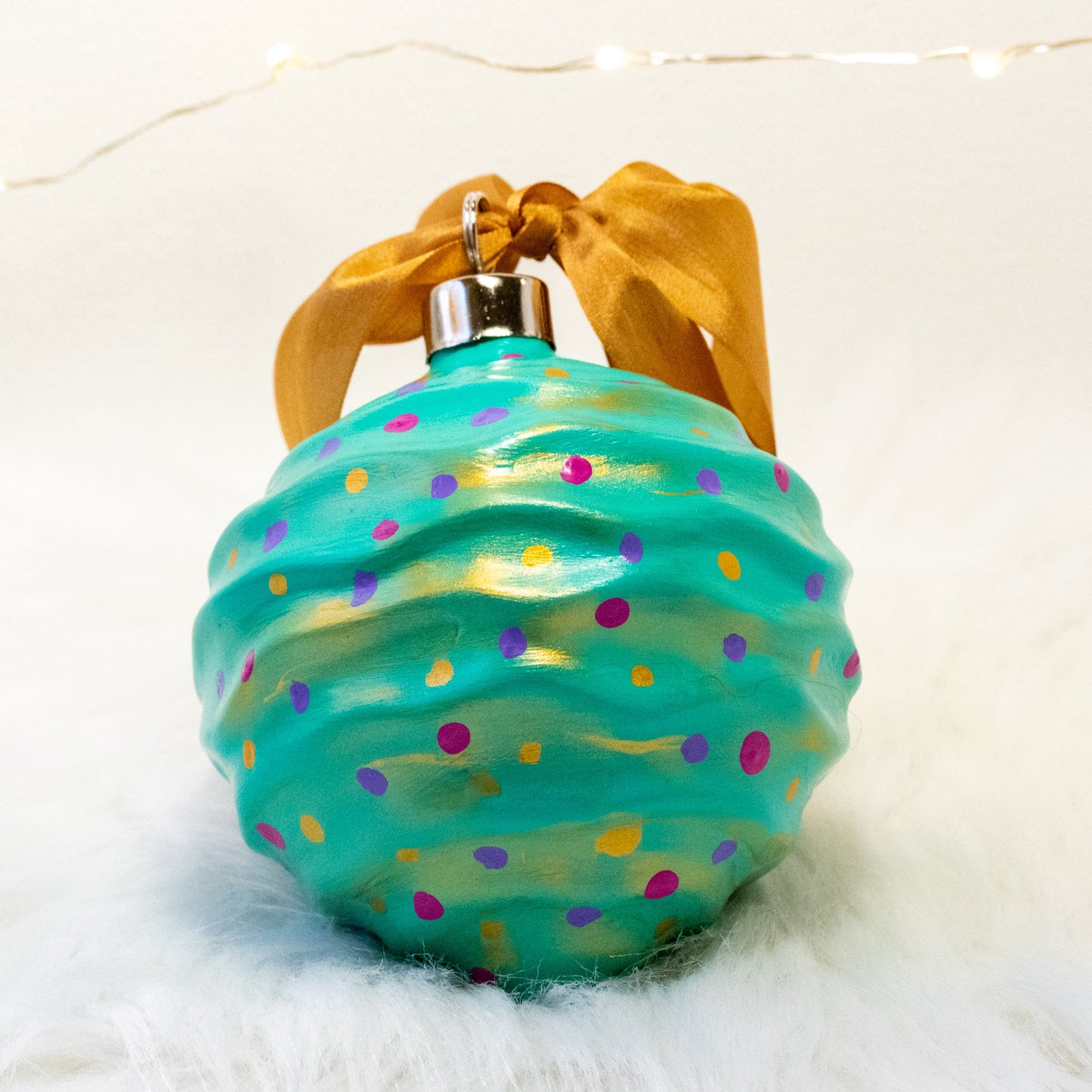 The Trina Hand Painted Ornament features a bright aqua green base coat with fuchsia, purple and gold polka dots and gold accents. Painted using fluid acrylic and acrylic paints. Displayed on white faux fur with fairy lights in the background.