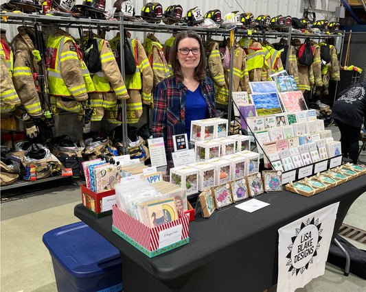 Photo of Lisa Blake standing behind her craft fair table filled with products.