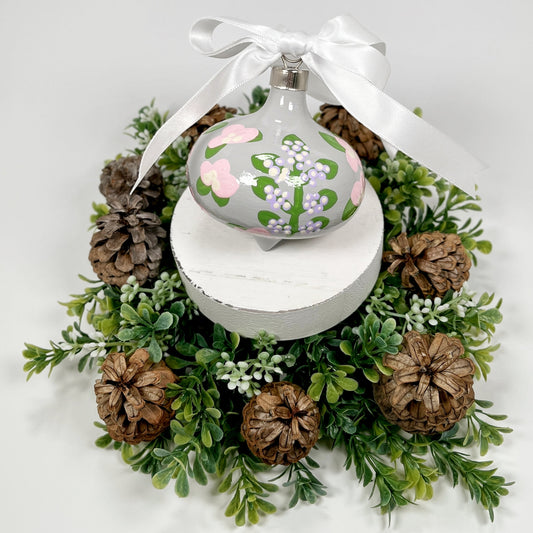 Decorative hand painted ornament with floral design on a white base surrounded by greenery and pinecones.