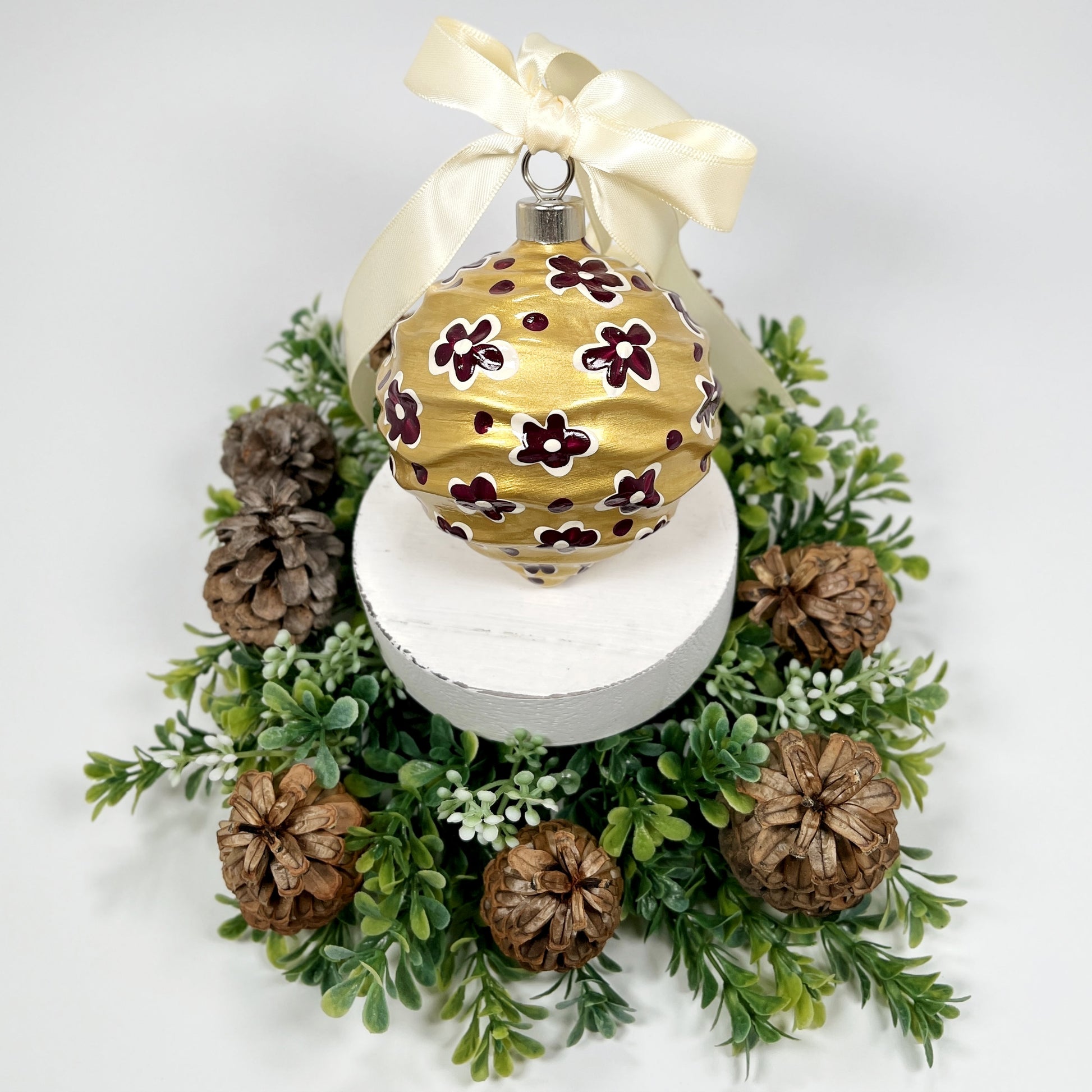 Decorative hand painted gold Christmas ornament with deep magenta and cream flowers and dots with a cream bow on a white base with greenery and pine cones.