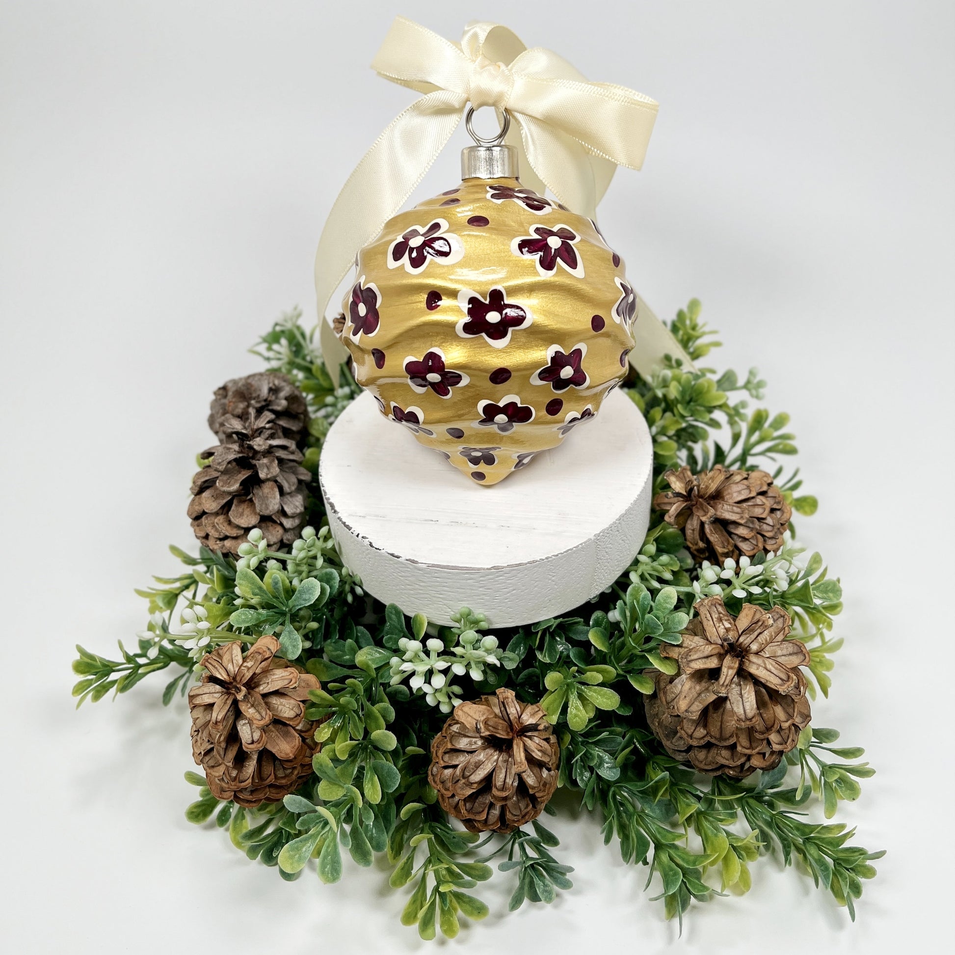 Decorative hand painted gold Christmas ornament with deep magenta and cream flowers and dots with a cream bow on a white base with greenery and pine cones.