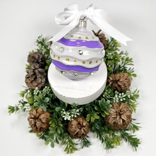 Decorative hand painted Christmas ornament with wavy purple stripes, gold polka dots and rhinestones with a white bow on a white base with greenery and pine cones.